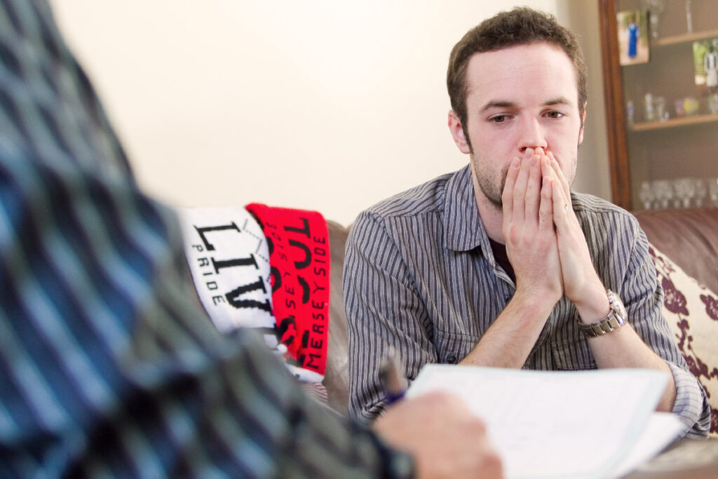 A man with his head in his hands sits on a sofa. He is watching another man fill in a form