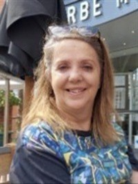 A white woman with long brown hair, wearing a blue patterned top, is smiling at the camera