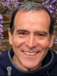 A white man with short dark hair and wearing a dark top is smiling at the camera