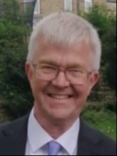 A white man with short grey hair and glasses, wearing a suit and tie, is smiling at the camera