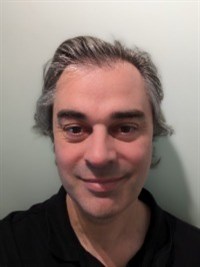 A white man with dark hair and wearing a black top is smiling at the camera