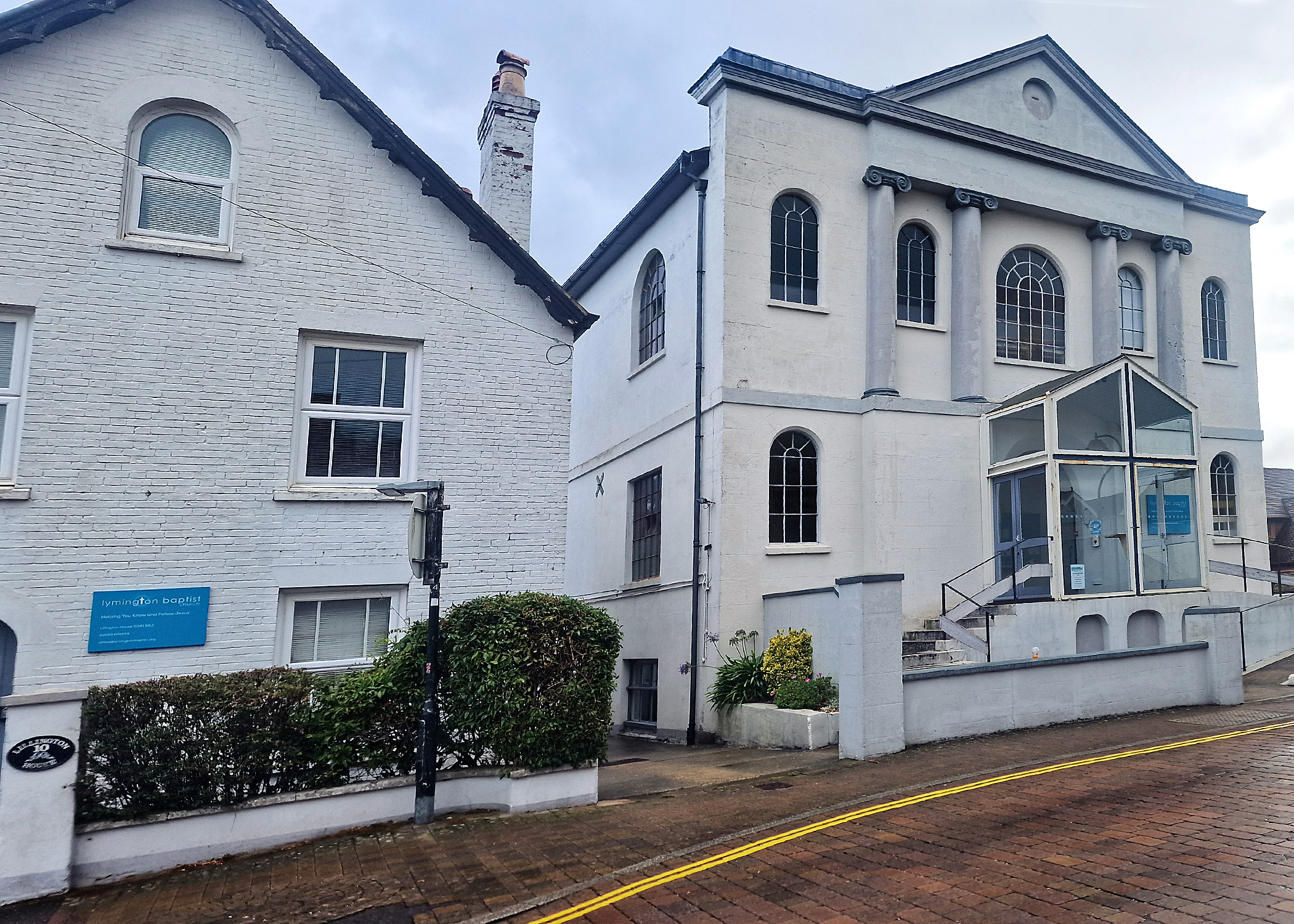 A white church building wuth stairs on the left and ramp on the right. There are double yellow lines on the road in front