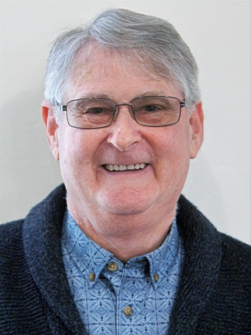 A white man with grey hair and glasses, wearing a blue shirt and cardigan smiling at the camera