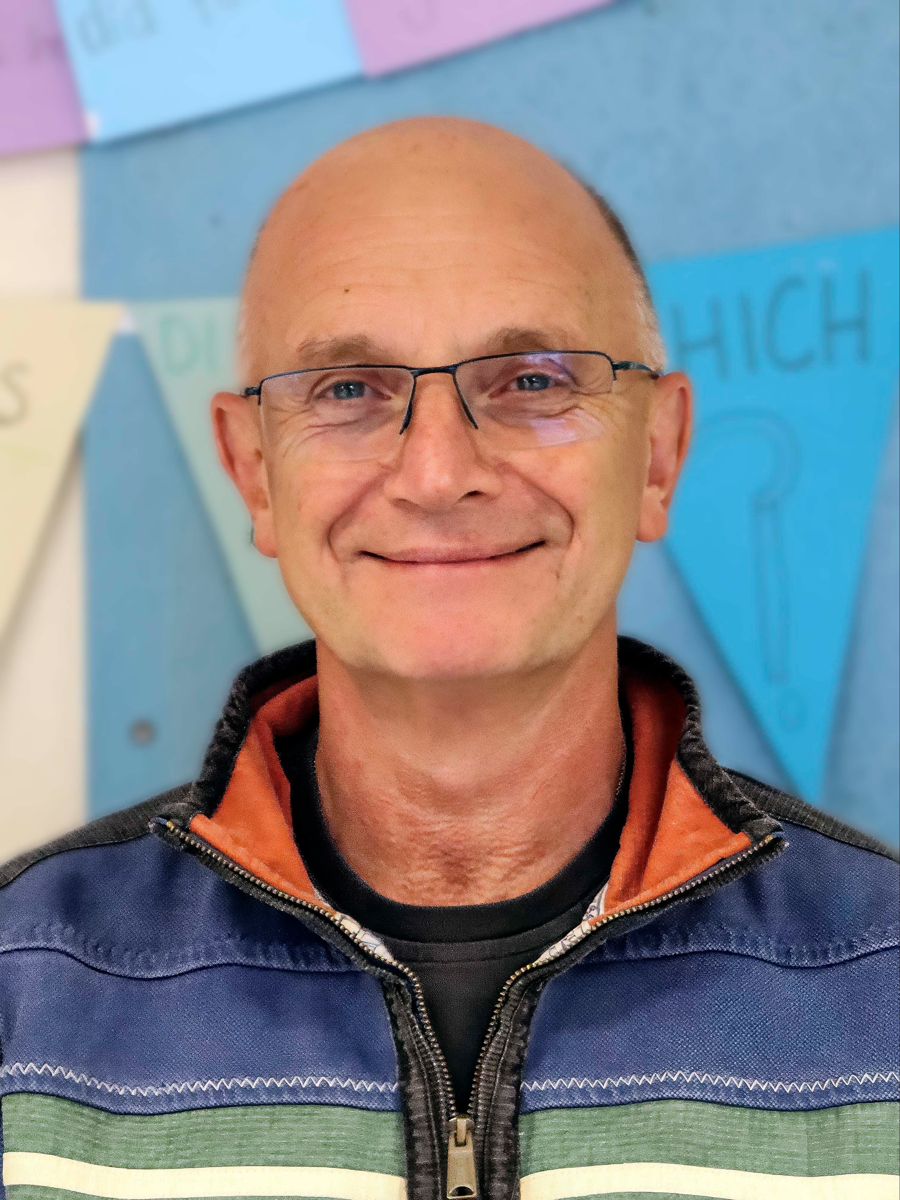 A white man with a bald head smiles at the camera. He is wearing glasses and a stripy top