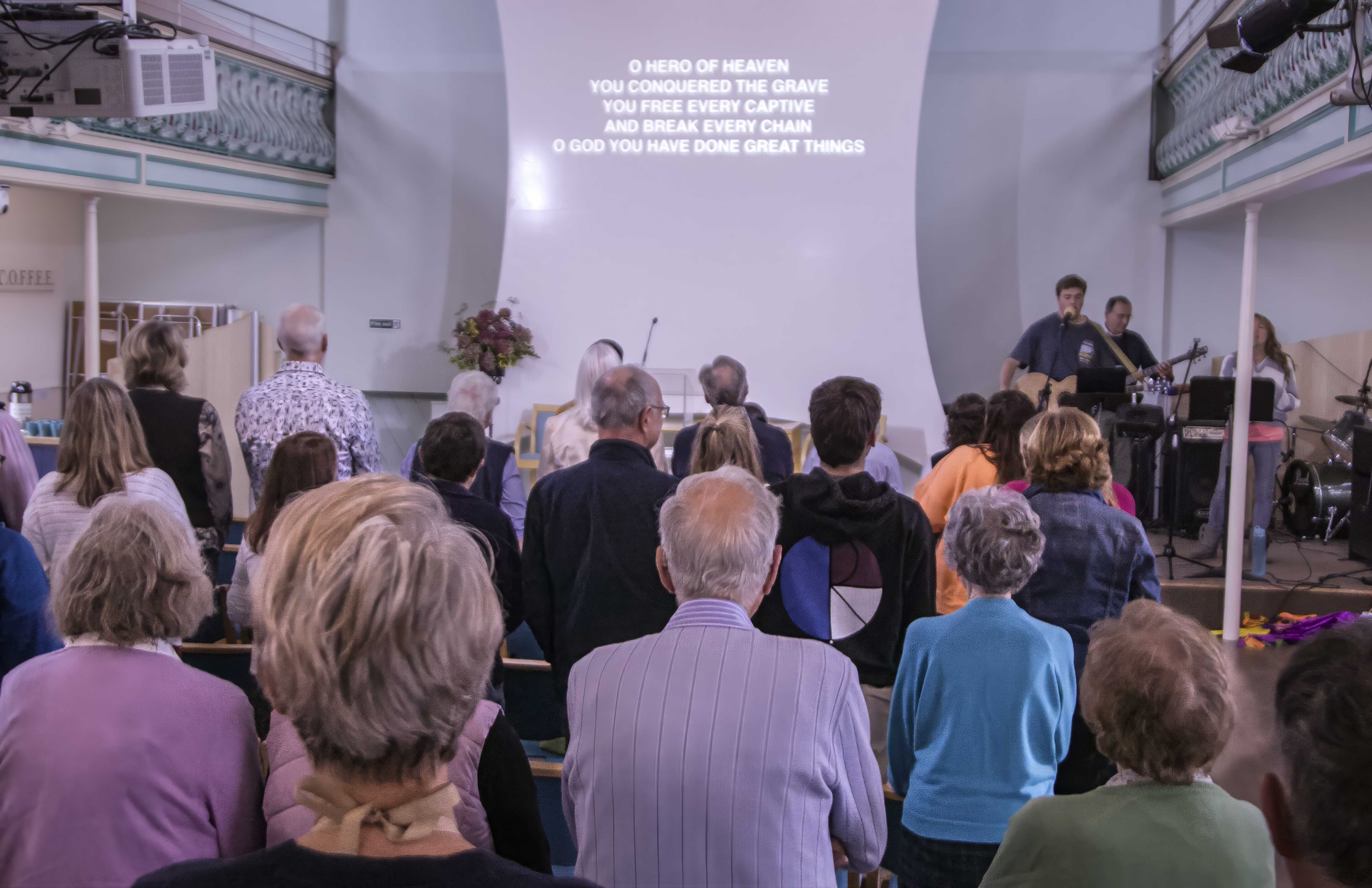 Band singing and playing on stage. Words of a song are projected onto a sail shaped screen. People standing in rows are facing the band