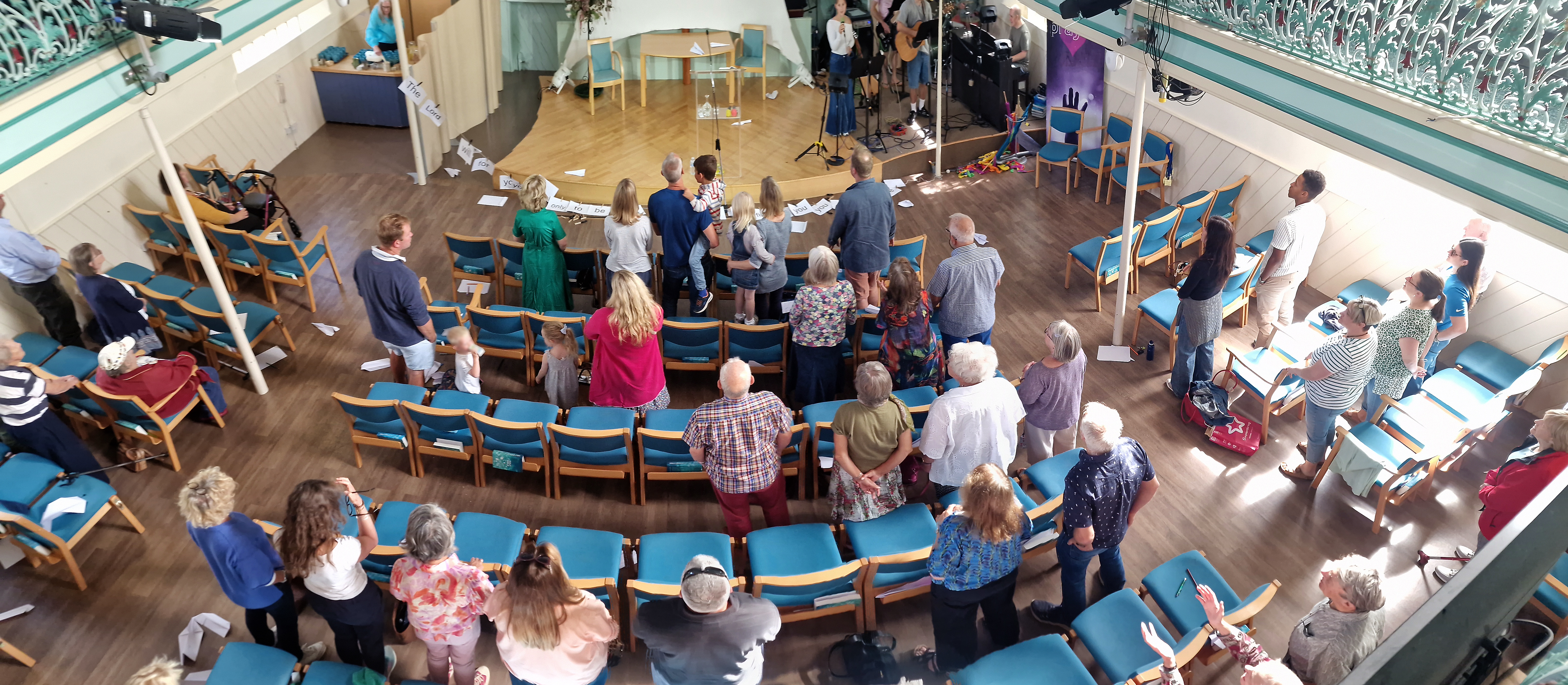 People are standing and singing. There is a worship band in the background. Photo is taken from over head 