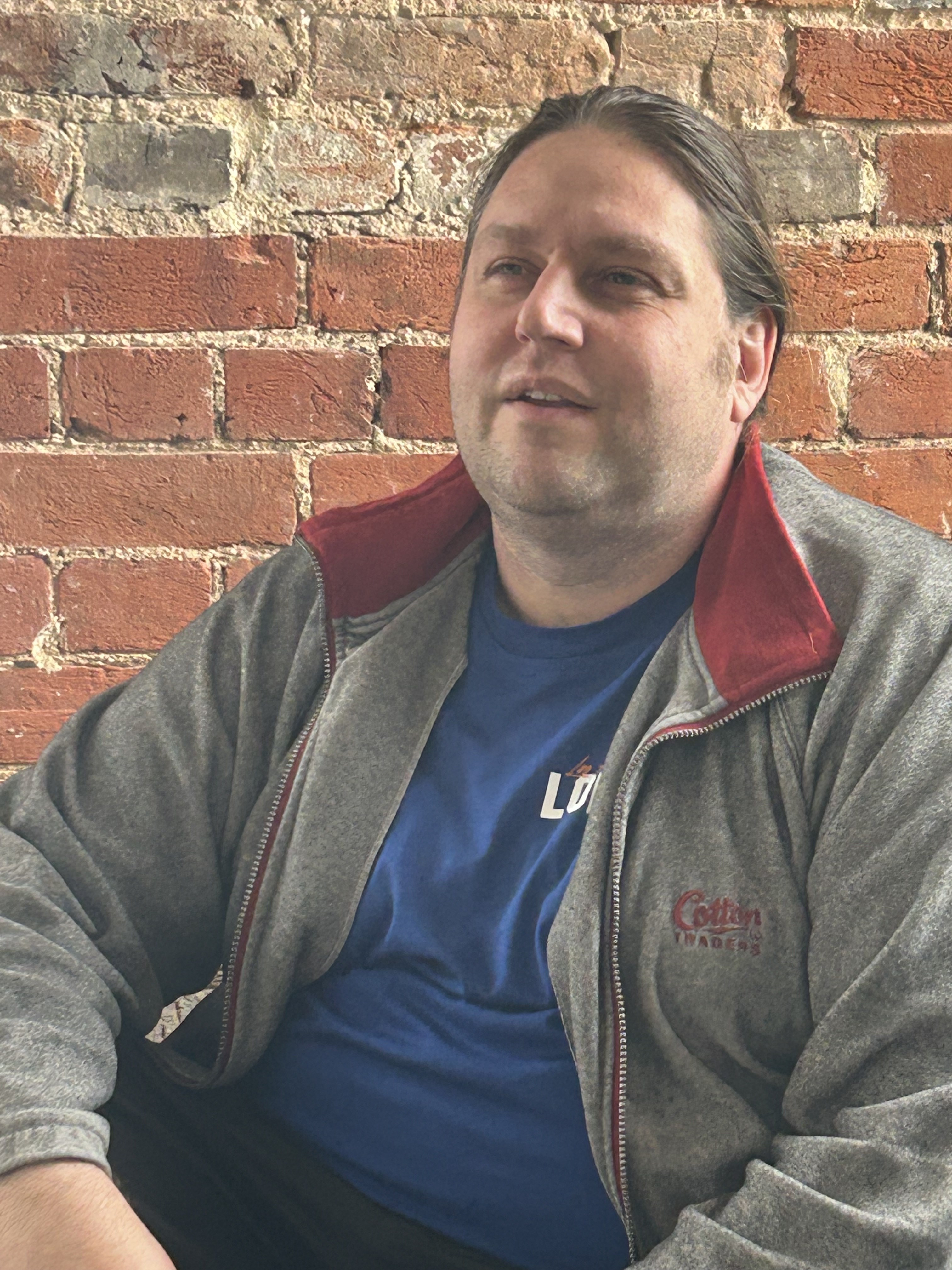 A white man with long brown hair in a ponytail looks off to the right. He's wearing a blue t-shirt and grey zipped jacket.
