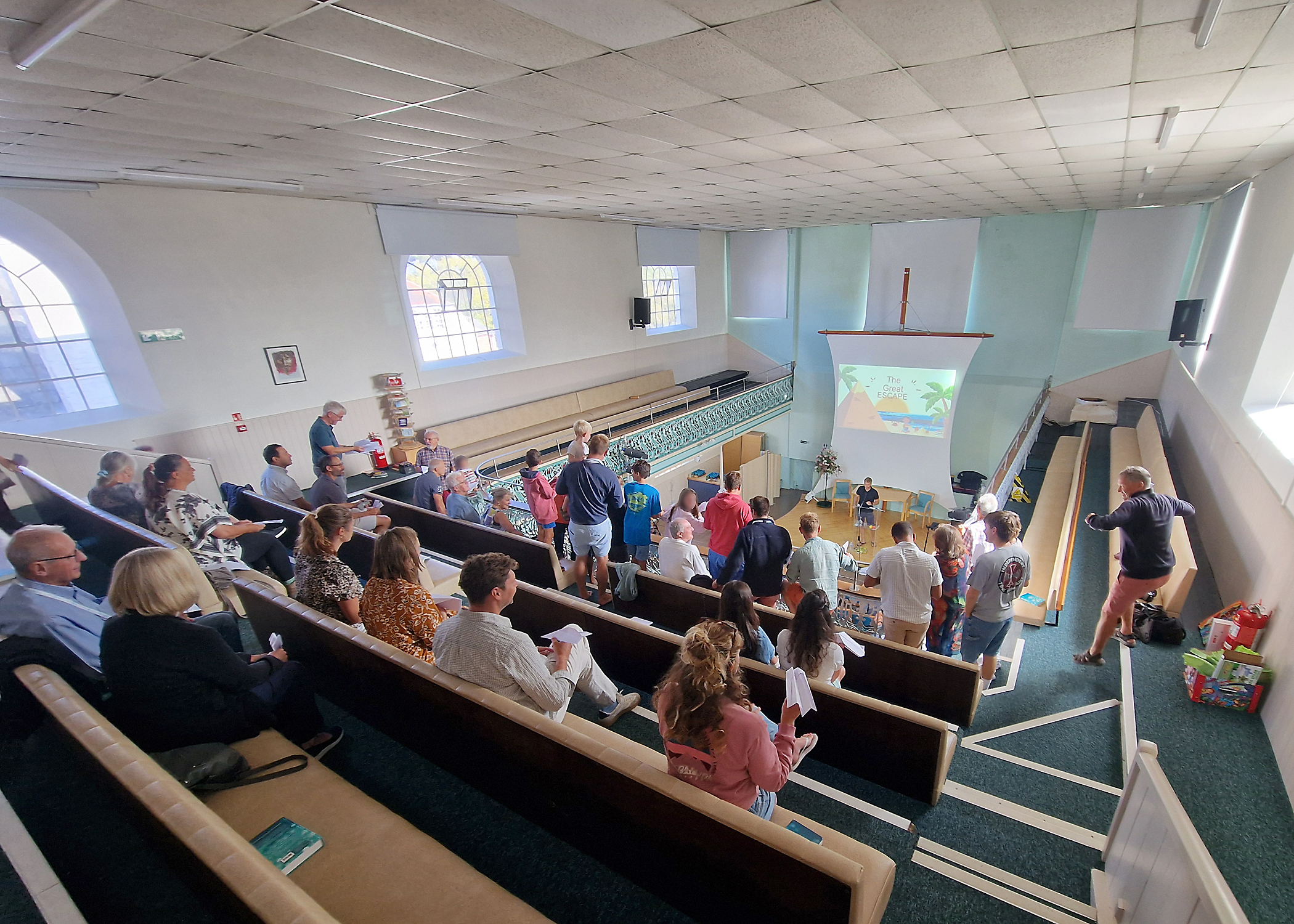 People seated and standing on the balcony. In the background there is someone on stage and a picture projected onto the sail shaped screen 