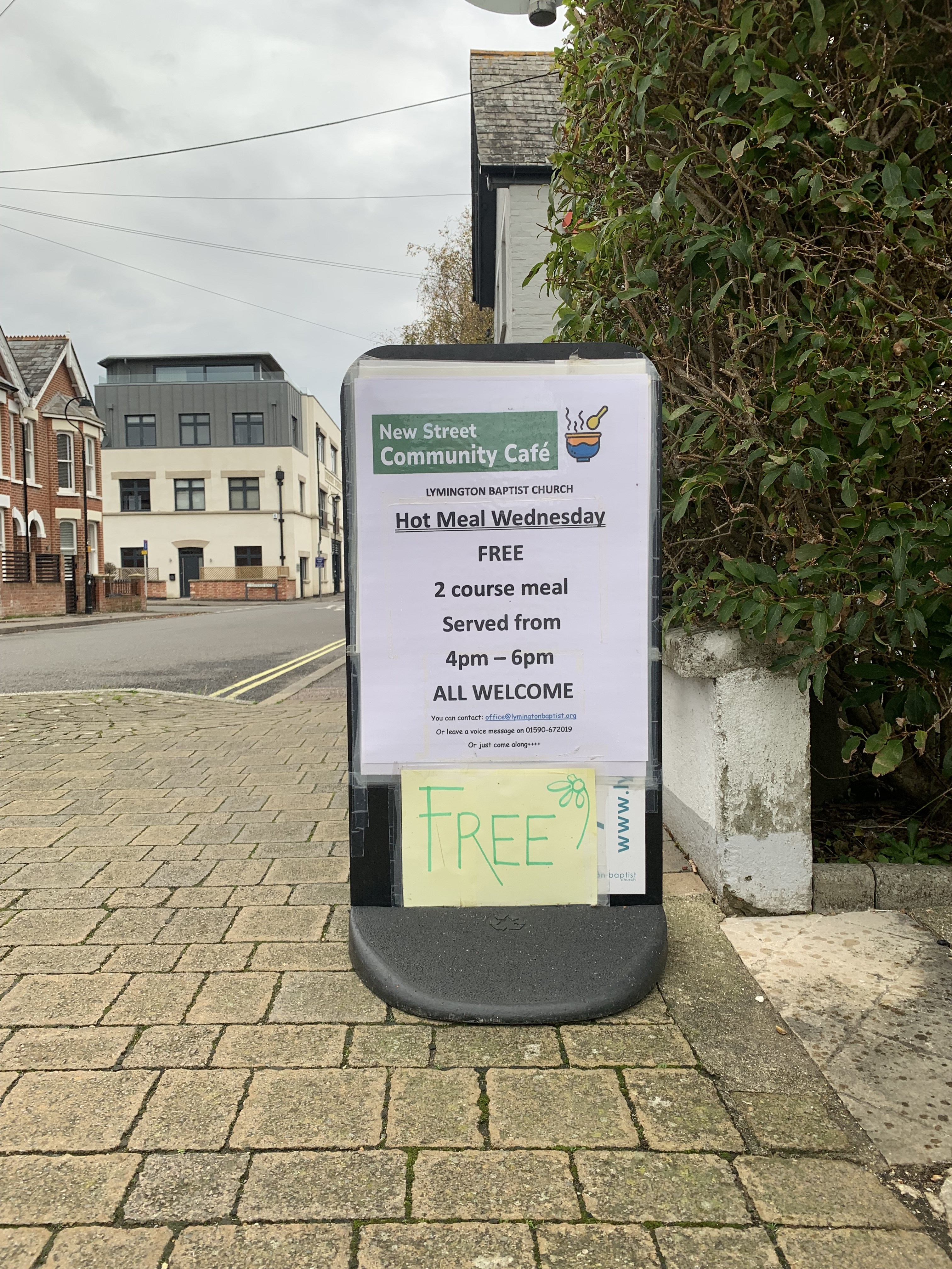 White street sign: Hot Meal Wednesday FREE. In the background is a view of the road. 