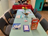 A table set with a green checked cloth, jenga, dominoes and playing cards. A table set with tea and coffee is in the background
