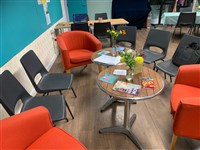 Orange and black chairs arranged in a circle around 2 coffee tables set with flyers and vases of yellow, orange and white flowers