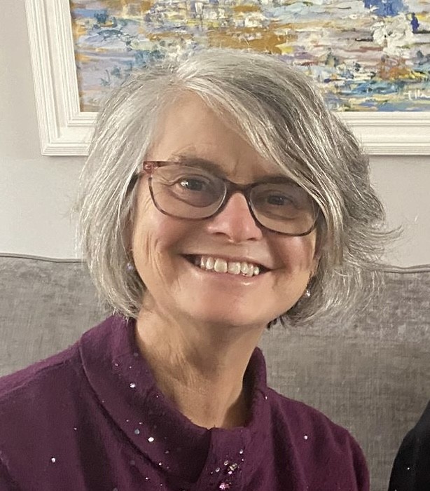 A woman with chin length grey hair and glasses, wearing a burgundy top, is smiling at the camera