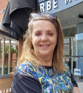 A white woman with long brown hair and a blue patterned top is smiling at the camera