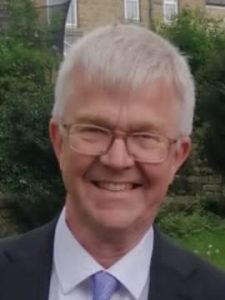 A white man with short grey hair and square framed glasses is smiling at the camera. He is wearing a suit and tie.