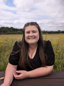 A white woman with long brown hair and a black top smiling at the camera. There is a green field behind her.