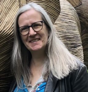 A white woman with long grey hair and glasses is smiling at the camera. She is standing next to a life size bamboo elephant.