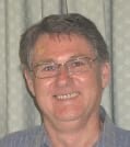 A white man with grey hair and glasses is smiling at the camera. He is wearing a grey shirt