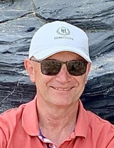 A white man wearing a white baseball cap, sunglasses and pink shirt smiling at the camera