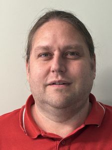 A white man with long brown hair in a pony tail and wearing a red polo shirt smiling at the camera